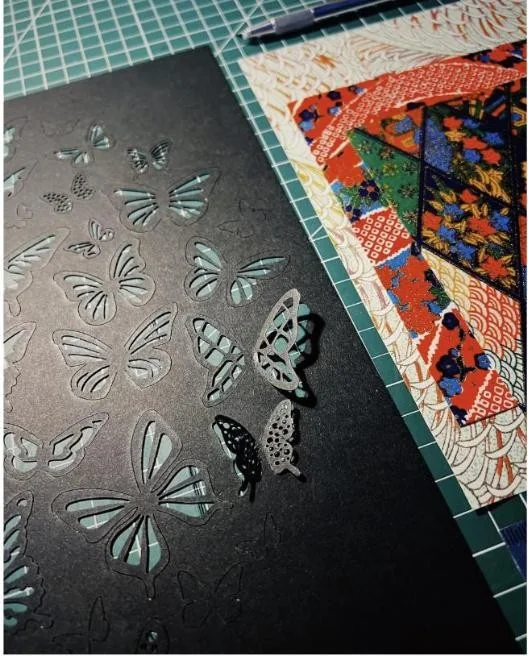 Laser-cut butterflies lifting out of black card beside patterned washi paper